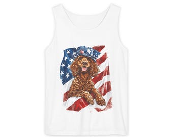 May include: White tank top featuring a brown dog wearing a patriotic hat, set against a backdrop of the American flag. The dog is depicted with a happy expression, and the flag's red, white, and blue colors are prominent.