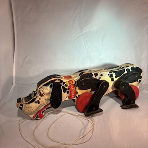 Op de afbeelding: Vintage trekdier speelgoedhond met zwarte vlekken, rode wielen en een rode halsband met de tekst "Snoopy". De hond heeft een crème kleur met zwarte vlekken. Het speelgoed heeft een touwtje om aan te trekken.