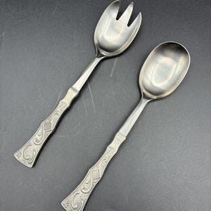 May include: A silver serving spoon and fork set. The fork has three tines. Both utensils have ornate handles with a diamond pattern and scrollwork. The set is displayed on a dark surface.