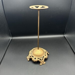 May include: A gold-toned metal display stand featuring a tall, slender vertical rod attached to a circular top plate with heart-shaped cutouts and a wide, ornate, decorative base with intricate scrollwork and a domed center.