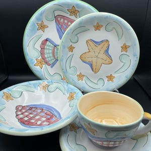 May include: A set of hand-painted ceramic tableware with a nautical theme. The set includes plates, bowls, and a teacup with a saucer. The designs feature seashells, starfish, and floral accents in shades of blue, red, and yellow.