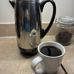 以下が含まれることがあります： 黒いハンドルとベースが付いたステンレス製のFarberwareコーヒーパーコレーターと、濃いコーヒーが入った白いマグカップ。コーヒー豆の入ったガラス瓶が背景にあります。