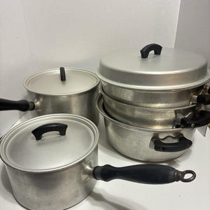 Op de afbeelding: Een verzameling vintage aluminium kookgerei, waaronder potten en een stoomkoker. De potten hebben zwarte handvatten en deksels met zwarte knoppen. Het kookgerei staat op een wit oppervlak.