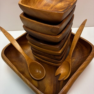 May include: A wooden salad serving set featuring a large square tray, a stack of square bowls, and two wooden serving spoons. The set is made of a warm-toned wood, showcasing the natural grain.