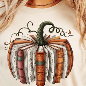 May include: Cream-colored t-shirt featuring a pumpkin design made of open books. The pumpkin has a green stem and vine-like tendrils. The books are orange, red, and brown, with white pages. A fall-themed graphic tee.