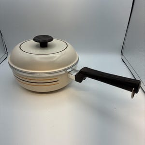 May include: A cream-coloured saucepan with a matching lid, featuring a dark brown knob and a long, dark brown handle. The exterior of the pan is decorated with two thin horizontal stripes in brown and gold, and a metal ring hangs from the handle.