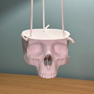 May include: White ceramic skull planter with a rope hanger. The planter is designed to hold small plants or flowers.