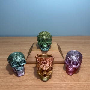 May include: Four decorative skull figurines in shades of green, gold, and purple are displayed on a wooden surface. The skulls have intricate designs and varying finishes. One skull is a container. The background is a solid blue wall.
