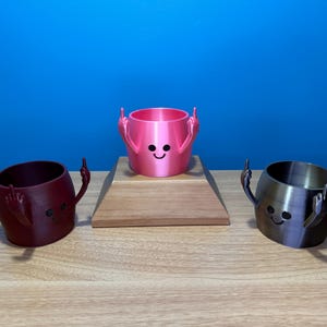 May include: Three 3D printed ceramic cups with a smiley face and two raised arms. The cups are pink, burgundy, and silver.