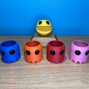 May include: Five Pac-Man ghost-shaped cups in blue, orange, red, pink, and yellow. The cups are arranged in a row on a wooden surface. A yellow Pac-Man figure is on a wooden platform behind the cups.
