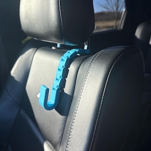 May include: A blue plastic car seat hook with a flexible design. The hook is attached to the headrest of a black leather car seat.