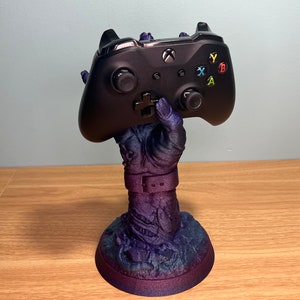 May include: A black Xbox controller rests on a purple and blue hand-shaped stand. The stand has a detailed, textured design, and the controller features colored buttons and the Xbox logo. The stand is on a wooden surface.