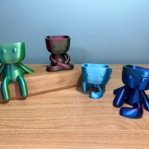 May include: Four 3D printed planters in the shape of a small person. The planters are in various colors, including green, blue, purple, and blue. The planters are sitting on a wooden surface.