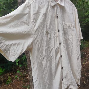May include: Cream-colored short-sleeved shirt with a button-down front and a chest pocket. The shirt is made of a lightweight fabric and has a relaxed fit. A small decorative element is sewn on the front.