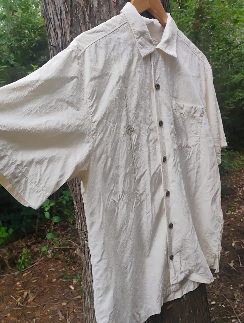 May include: Cream-colored short-sleeved shirt with a button-down front and a chest pocket. The shirt is made of a lightweight fabric and has a relaxed fit. A small decorative element is sewn on the front.
