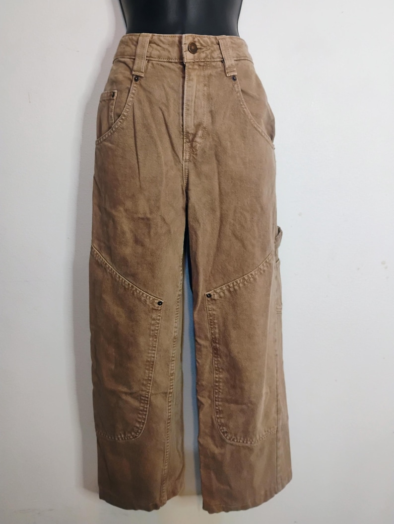 May include: High-waisted tan utility pants featuring a button closure, belt loops, and reinforced knee panels. These pants include deep front pockets with metal rivets and a side utility loop, constructed from a durable, solid-colored cotton fabric.