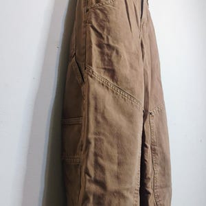 May include: A pair of brown utility pants featuring a high-waisted design, a button and zipper fly closure, and reinforced panels on the front legs. These pants include side pockets and visible stitching details along the seams and leg panels.