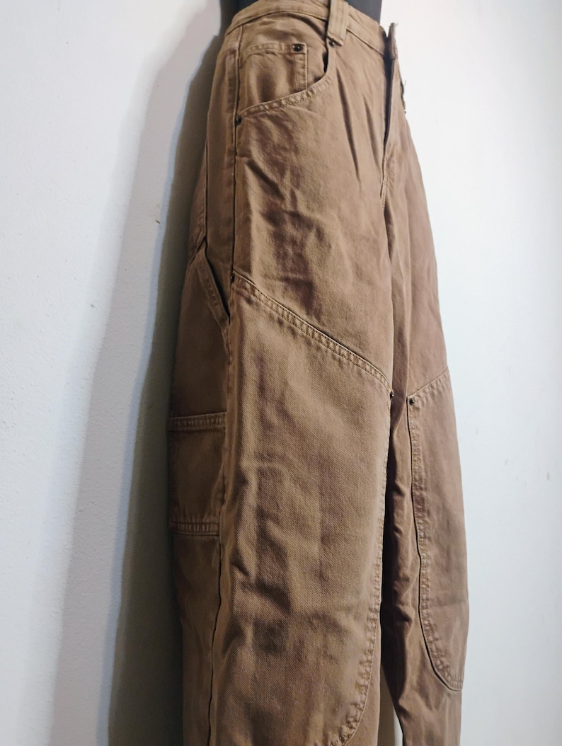 May include: A pair of brown utility pants featuring a high-waisted design, a button and zipper fly closure, and reinforced panels on the front legs. These pants include side pockets and visible stitching details along the seams and leg panels.
