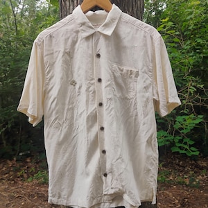 May include: Cream-colored short-sleeved button-down shirt with a single chest pocket and brown buttons. The shirt is hanging on a wooden hanger. A small decorative element is stitched on the left side of the shirt.