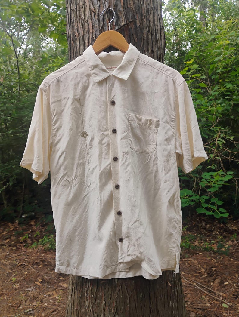 May include: Cream-colored short-sleeved button-down shirt with a single chest pocket and brown buttons. The shirt is hanging on a wooden hanger. A small decorative element is stitched on the left side of the shirt.