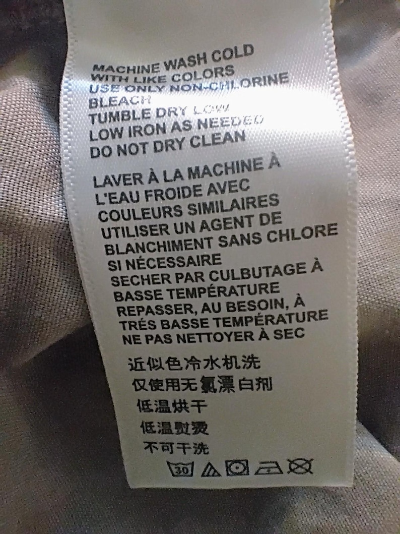 May include: A white fabric care label with black text in English, French, and Chinese. The instructions read: MACHINE WASH COLD WITH LIKE COLORS, USE ONLY NON-CHLORINE BLEACH, TUMBLE DRY LOW, LOW IRON AS NEEDED, DO NOT DRY CLEAN, and laundry symbols.