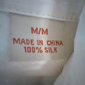 May include: Close-up of a clothing tag with the text "M/M MADE IN CHINA 100% SILK" in orange lettering on a beige background. The tag is sewn into a white fabric, likely a shirt or blouse.