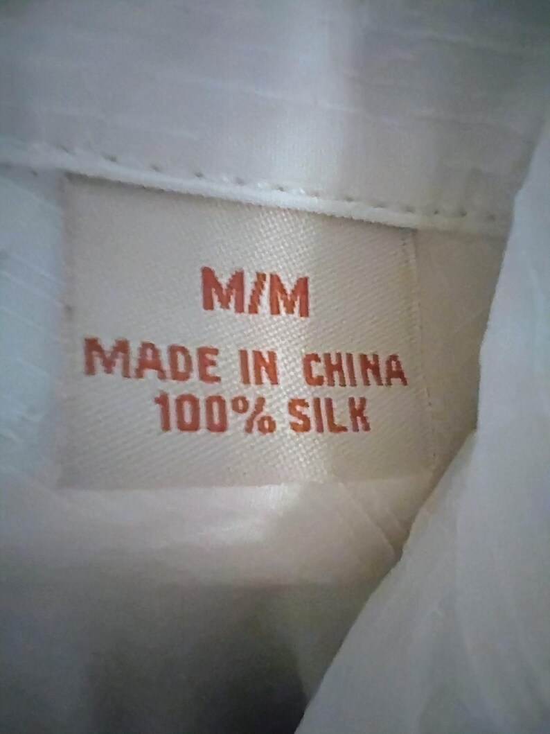 May include: Close-up of a clothing tag with the text "M/M MADE IN CHINA 100% SILK" in orange lettering on a beige background. The tag is sewn into a white fabric, likely a shirt or blouse.