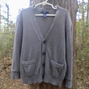 May include: A gray cardigan sweater with a V-neck, two front pockets, and button closures. The sweater is hanging on a white hanger against a tree trunk background. The Gap brand label is visible on the hanger.