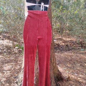 May include: A pair of high-waisted, flared pants in a deep red hue. The pants have a vertical ribbed texture and are displayed on a mannequin. A white, crocheted halter top is also visible. The background features a natural outdoor setting.