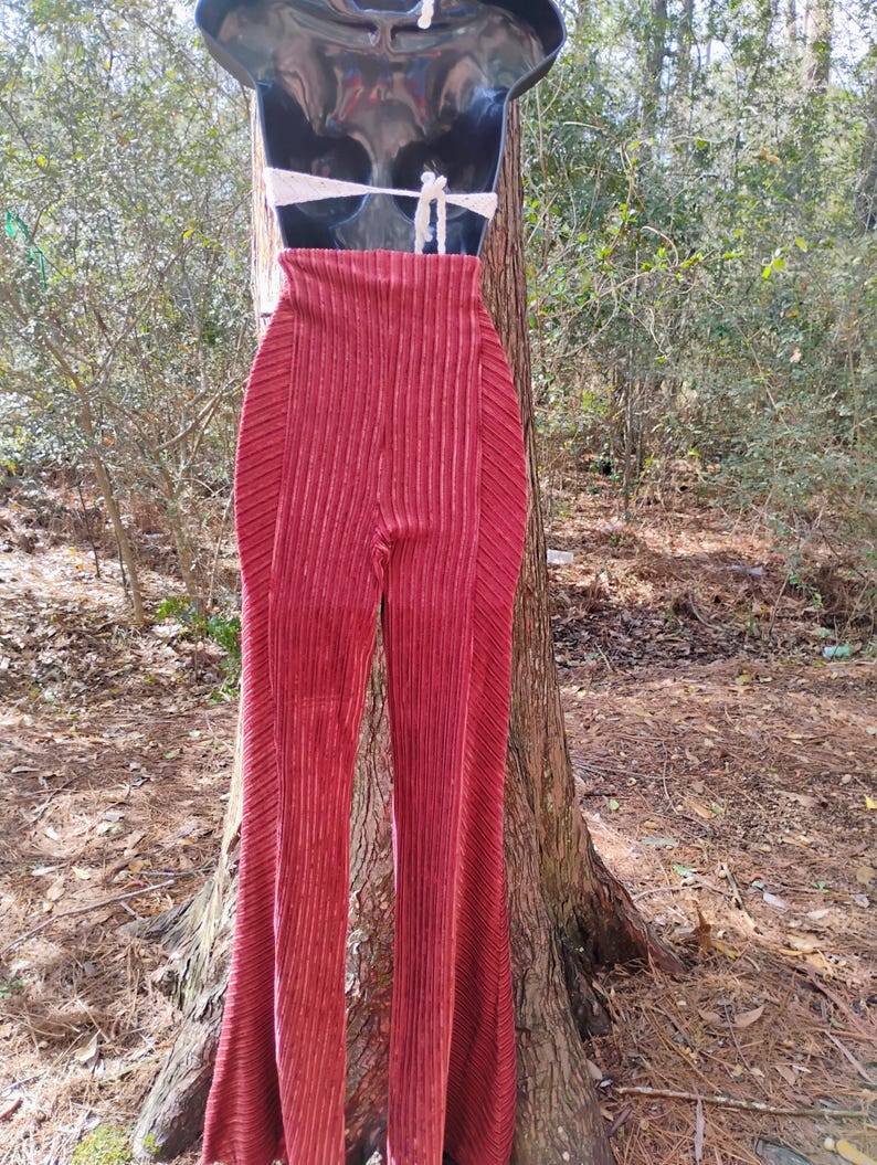 May include: A pair of high-waisted, flared pants in a deep red hue. The pants have a vertical ribbed texture and are displayed on a mannequin. A white, crocheted halter top is also visible. The background features a natural outdoor setting.