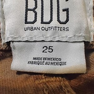 May include: A white fabric clothing tag with black text reading BDG URBAN OUTFITTERS, the number 25, and the text MADE IN MEXICO FABRIQU&Eacute; AU MEXIQUE. The tag is stitched onto a brown textured garment with visible seams and fabric weave details.