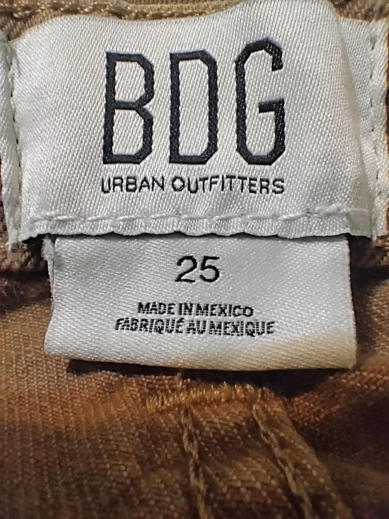 May include: A white fabric clothing tag with black text reading BDG URBAN OUTFITTERS, the number 25, and the text MADE IN MEXICO FABRIQU&Eacute; AU MEXIQUE. The tag is stitched onto a brown textured garment with visible seams and fabric weave details.