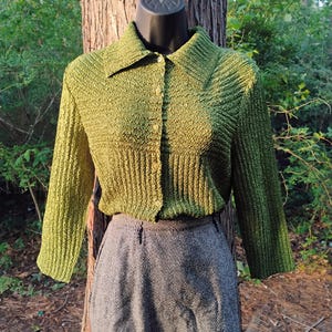 May include: A green textured button-down shirt with long sleeves and a collar, paired with a gray and black patterned skirt. The shirt is buttoned up and displayed on a mannequin. The background features a tree trunk and foliage.