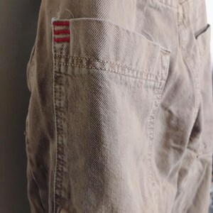 May include: A pair of tan denim pants featuring a large patch pocket on the side. The pocket is accented with three horizontal red embroidered lines at the top corner. The fabric has a visible twill weave texture and reinforced stitching throughout.