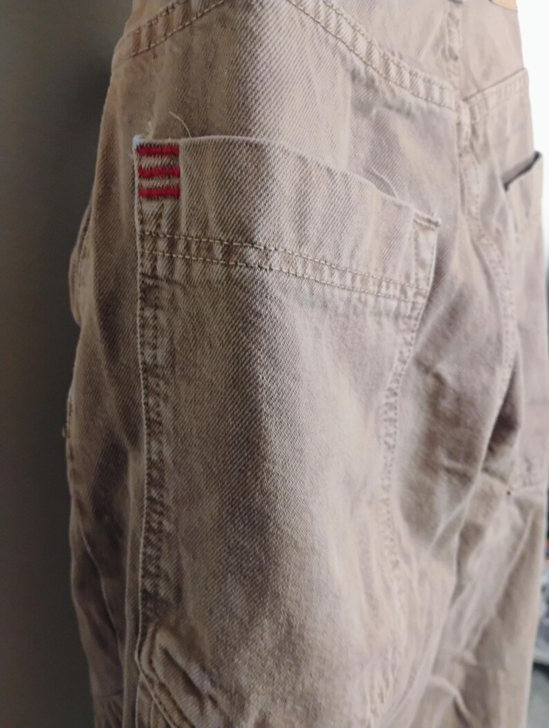 May include: A pair of tan denim pants featuring a large patch pocket on the side. The pocket is accented with three horizontal red embroidered lines at the top corner. The fabric has a visible twill weave texture and reinforced stitching throughout.