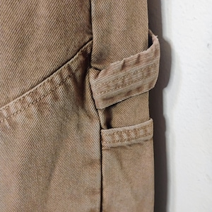 May include: A pair of tan cotton twill pants featuring a reinforced side utility loop and a deep diagonal pocket. The fabric displays a distinct diagonal weave pattern with heavy-duty double-stitched seams along the pocket edge and the loop attachment.