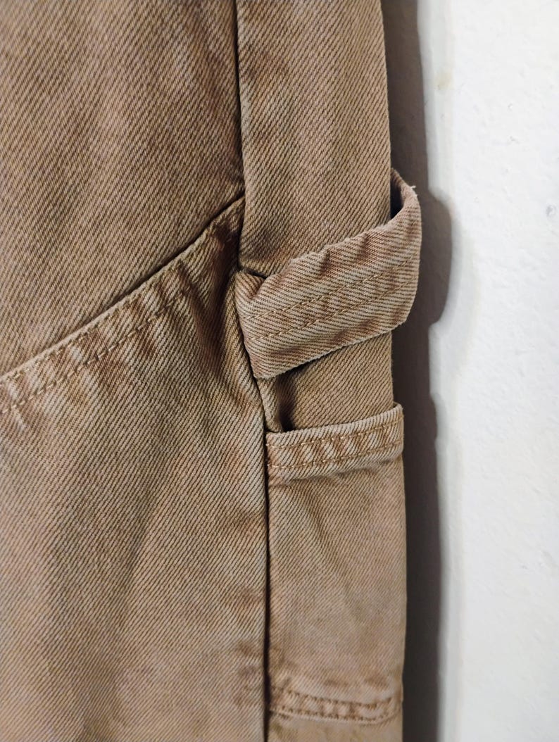 May include: A pair of tan cotton twill pants featuring a reinforced side utility loop and a deep diagonal pocket. The fabric displays a distinct diagonal weave pattern with heavy-duty double-stitched seams along the pocket edge and the loop attachment.