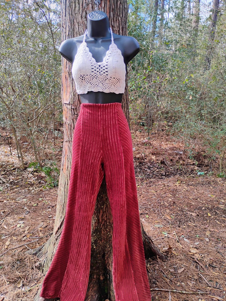 May include: A white crochet halter top and high-waisted, wide-leg, burgundy velvet pants displayed on a mannequin. The pants have vertical pleats. The outfit is displayed outdoors against a tree trunk.