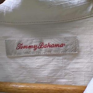 May include: Close-up of a white button-down shirt with a textured pattern. The shirt is hanging on a wooden hanger. The label reads "Tommy Bahama" in red script.