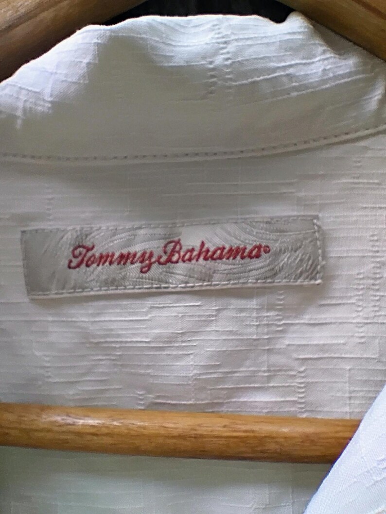 May include: Close-up of a white button-down shirt with a textured pattern. The shirt is hanging on a wooden hanger. The label reads "Tommy Bahama" in red script.