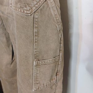 May include: Tan-colored utility pants featuring a prominent side pocket with reinforced stitching and a deep angled hip pocket. The fabric has a visible twill weave texture, and the garment includes a structured waistband with a top-stitched seam.
