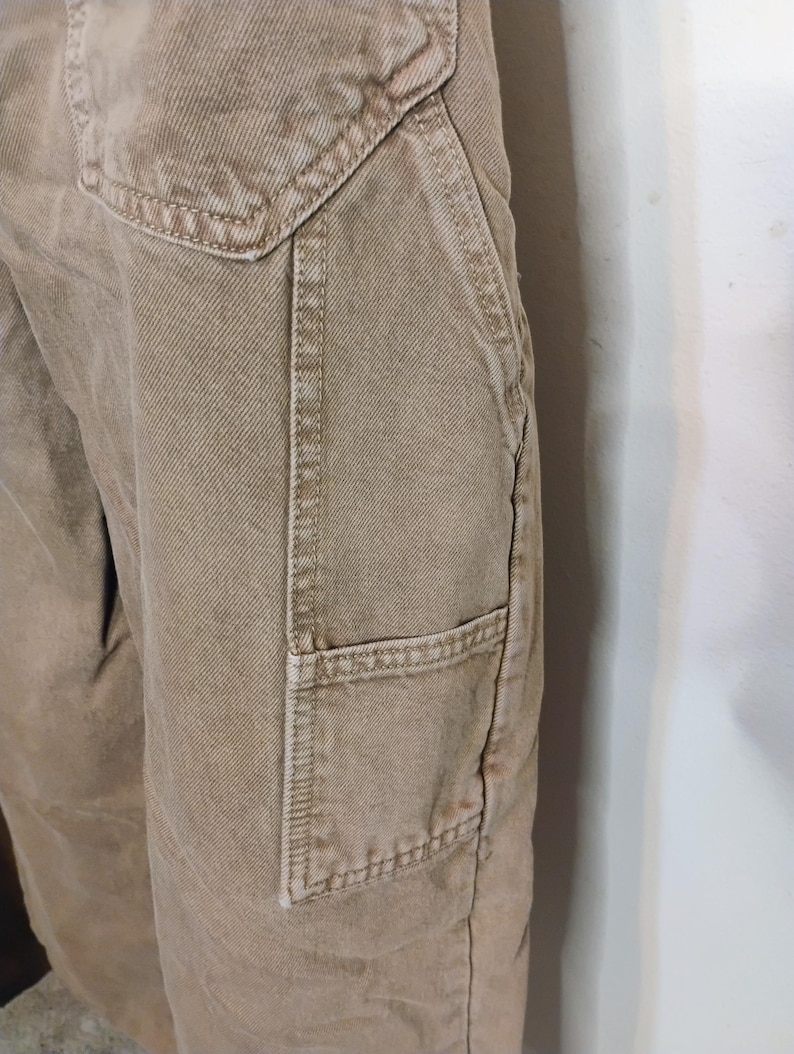 May include: Tan-colored utility pants featuring a prominent side pocket with reinforced stitching and a deep angled hip pocket. The fabric has a visible twill weave texture, and the garment includes a structured waistband with a top-stitched seam.