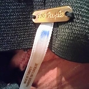 May include: Close-up of a gold-colored metal tag with the words "Free People" in cursive script. A white fabric tag with blue ink stains is attached. The fabric tag reads "MADE IN TURKEY FABRIQUE EN TURQUIE".