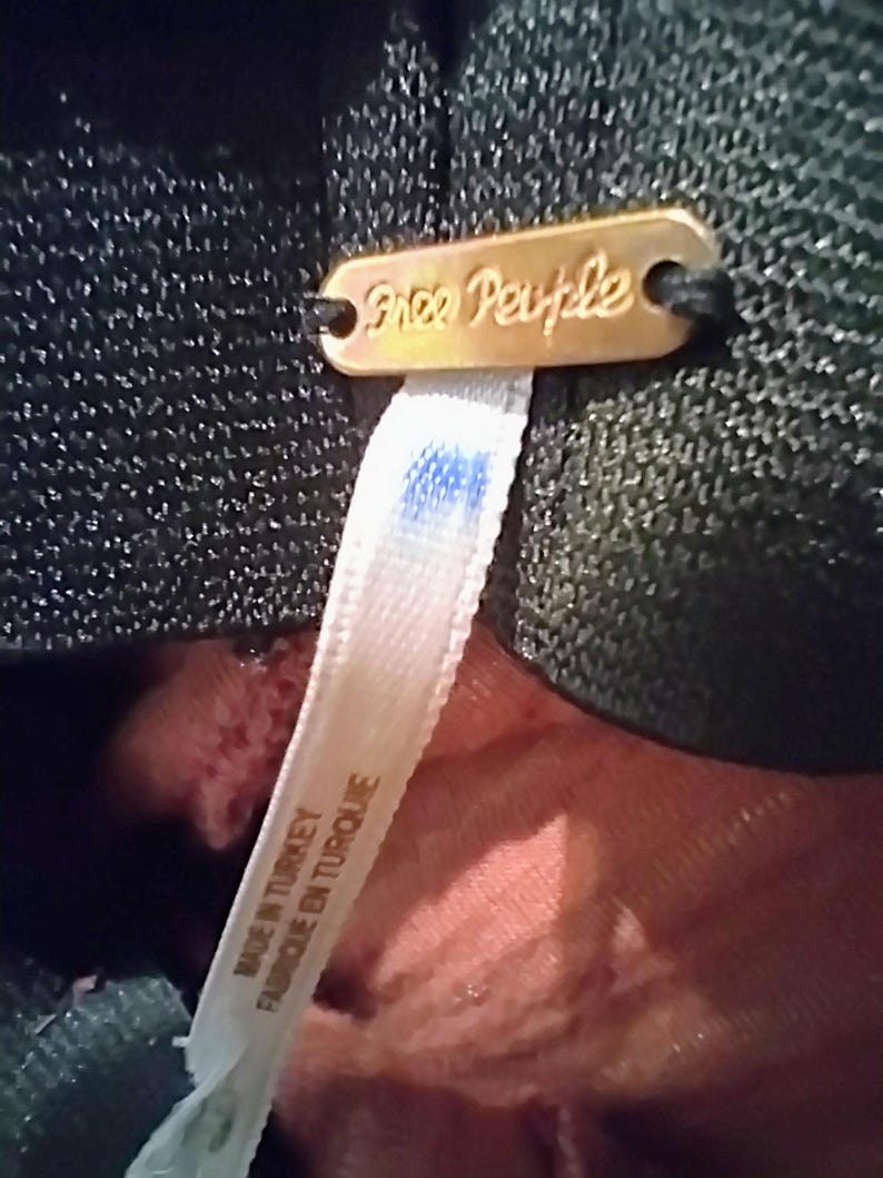 May include: Close-up of a gold-colored metal tag with the words "Free People" in cursive script. A white fabric tag with blue ink stains is attached. The fabric tag reads "MADE IN TURKEY FABRIQUE EN TURQUIE".