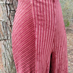 May include: Close-up of a pair of high-waisted, wide-leg pants in a deep rose color. The pants feature a vertical ribbed texture on one leg and a diagonal ribbed texture on the other, creating a unique design. The fabric appears to be soft and flowing.