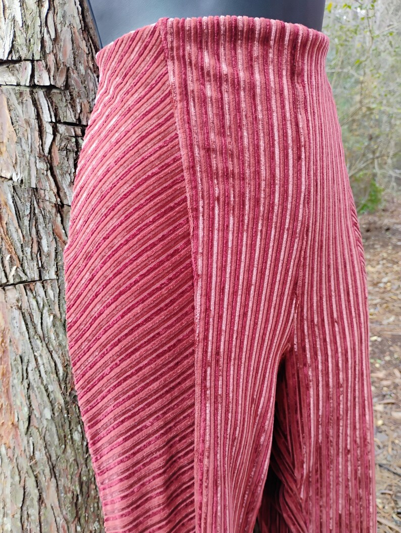 May include: Close-up of a pair of high-waisted, wide-leg pants in a deep rose color. The pants feature a vertical ribbed texture on one leg and a diagonal ribbed texture on the other, creating a unique design. The fabric appears to be soft and flowing.