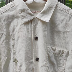 May include: Cream-colored button-down shirt with a single pocket and decorative embroidery. The shirt has a collared neckline and is hanging on a wooden hanger. The brand name "Tommy Bahama" is visible on the collar.