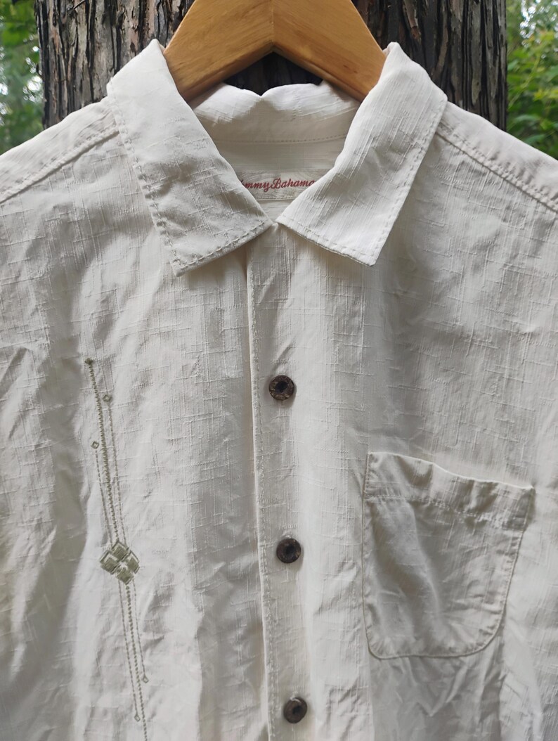 May include: Cream-colored button-down shirt with a single pocket and decorative embroidery. The shirt has a collared neckline and is hanging on a wooden hanger. The brand name "Tommy Bahama" is visible on the collar.