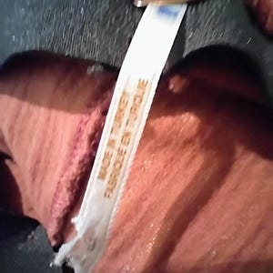 May include: Close-up of a tag that reads "MADE IN TURKEY FABRIQUE EN TURQUIE" in white text. The tag is attached to a reddish-orange fabric. A small, gold-colored metal piece is visible at the top of the image.