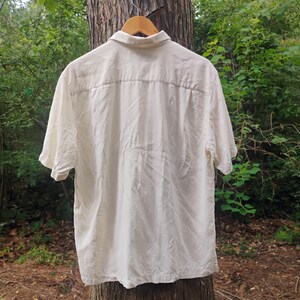 May include: Cream-colored short-sleeve shirt hanging on a wooden hanger. The shirt has a button-down front and a collar. The back of the shirt is visible, showing a yoke detail. The shirt is made of a lightweight fabric.
